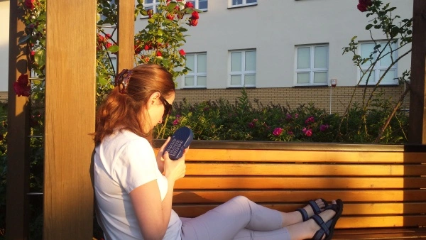 Kobieta w przeciwsłonecznych okularach siedząca na ławce w ogrodzie kwiatowym trzyma w dłoniach odtwarzacz audiobooków Czytak 4.